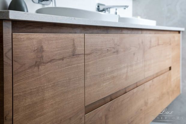 Close-up of a contemporary bathroom vanity with warm wood-grain cabinetry, integrated recessed drawer pulls, a pale stone benchtop, white vessel basins, and chrome tapware. A light grey textured wall finish is visible behind the vanity.