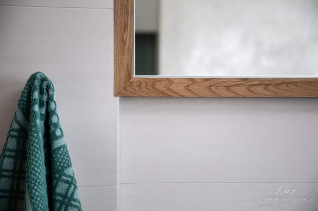 Close-up of a softly lit bathroom wall with large pale grey rectangular tiles, a timber-framed wall mirror, and a textured teal-green towel hanging at the left edge. The composition highlights the tile layout, natural wood grain, and restrained contemporary detailing.