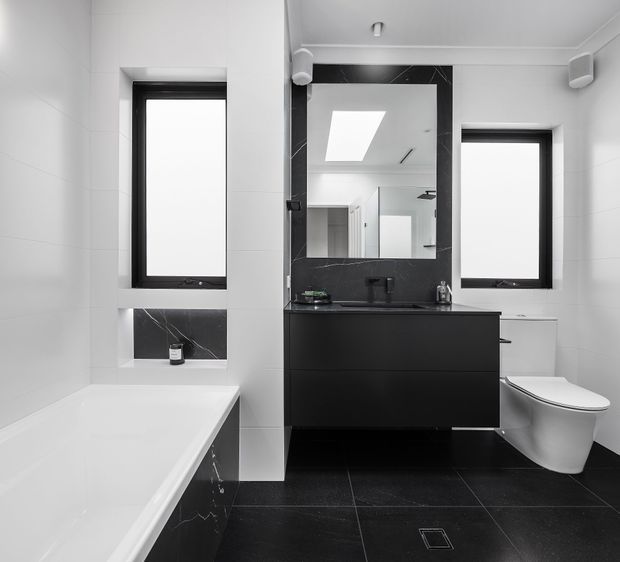 Contemporary monochrome bathroom featuring a white bathtub, black tiled floor, black marble-look wall accents, a floating black vanity with integrated basin, large mirror, wall-mounted speakers, black-framed windows, and a white toilet.