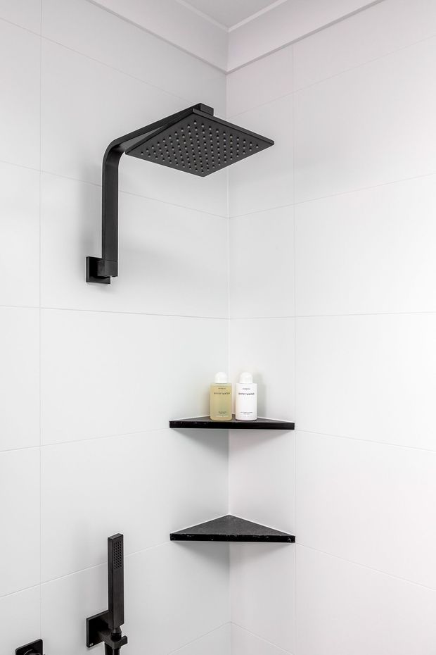 A minimalist walk-in shower enclosure with large white wall tiles, matte black rainfall showerhead and hand shower, and black triangular corner shelves holding toiletries. The space is bright, clean, and contemporary with strong black-and-white contrast.