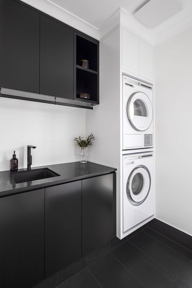 Compact contemporary laundry room with matte black cabinetry, dark benchtop and integrated black sink, white tiled walls, stacked white washer and dryer, and dark floor tiles. The space is neat, highly functional, and strongly monochromatic with black and white finishes.