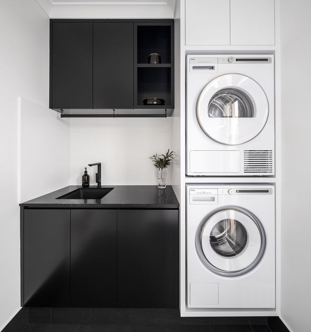Compact contemporary laundry room with stacked white washer and dryer integrated beside matte black cabinetry. The room features a dark stone-look benchtop with inset sink and black tapware, white wall finishes, overhead black storage, and open shelving with minimal decor.