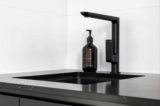 Close-up of a sleek black kitchen sink and high-arched black pull-out tap set into a dark benchtop, with a matching dark soap dispenser. The sink area is framed against white tiled walls in a minimalist, high-contrast contemporary fit-out.