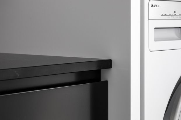Close-up monochrome detail of a sleek black built-in or freestanding unit with a narrow recessed opening, positioned beside a white Asko Quattro Conform washing machine or dryer. The surrounding wall is smooth grey, creating a minimalist, high-contrast composition.