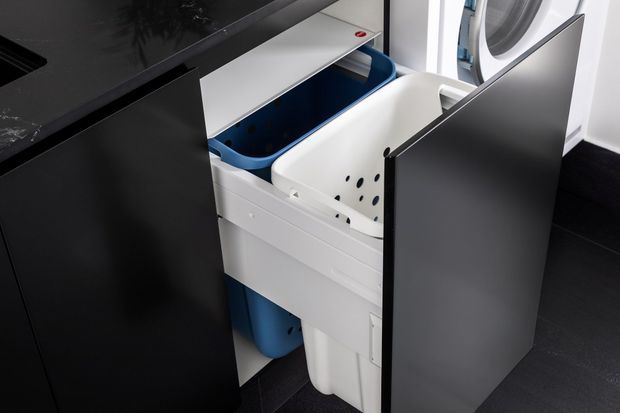 Close-up view of a compact laundry fit-out with a front-loading washing machine and pull-out laundry hampers or sorting bins housed in dark cabinetry. The cabinetry has a sleek matte finish, with white and blue plastic laundry baskets visible inside. Dark floor and countertop surfaces create a high-contrast contemporary utility space.