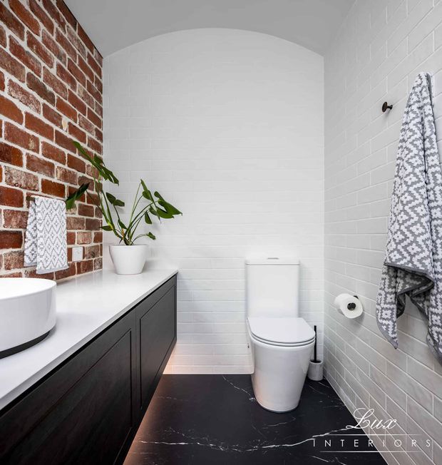 A compact contemporary bathroom with a dark floating vanity and white vessel basin, white subway-tiled walls, exposed red brick feature wall, black marble-look floor tiles, and a white toilet. A potted plant, patterned towels, and warm under-vanity lighting add contrast and texture.
