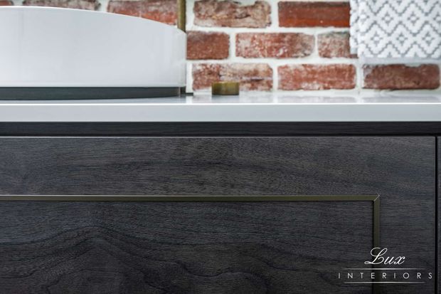 Close-up detail of dark textured timber cabinetry with slim metallic edging and a light benchtop. A red brick splashback forms the background, with a white appliance partially visible above the cabinetry. The image emphasizes material, grain, and joinery detailing.