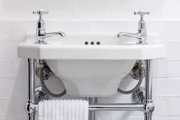 Close-up of a traditional-style white ceramic wall-mounted bathroom basin with twin chrome cross-handle taps, exposed polished chrome pipework, and a towel rail. The basin is set against a white subway-tile wall.