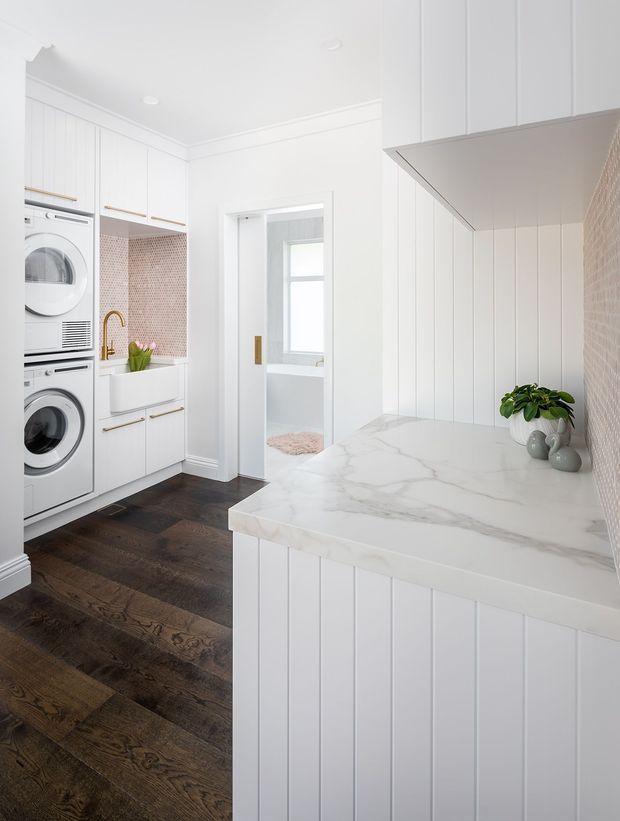 Bright, compact white laundry room with stacked front-loading washer and dryer, extensive white shaker-style cabinetry, vertical panelled cabinetry, a large white marble-look benchtop, pale pink mosaic tile splashback, dark timber flooring, and a small utility sink with brass tap. A doorway opens to a bathroom beyond.