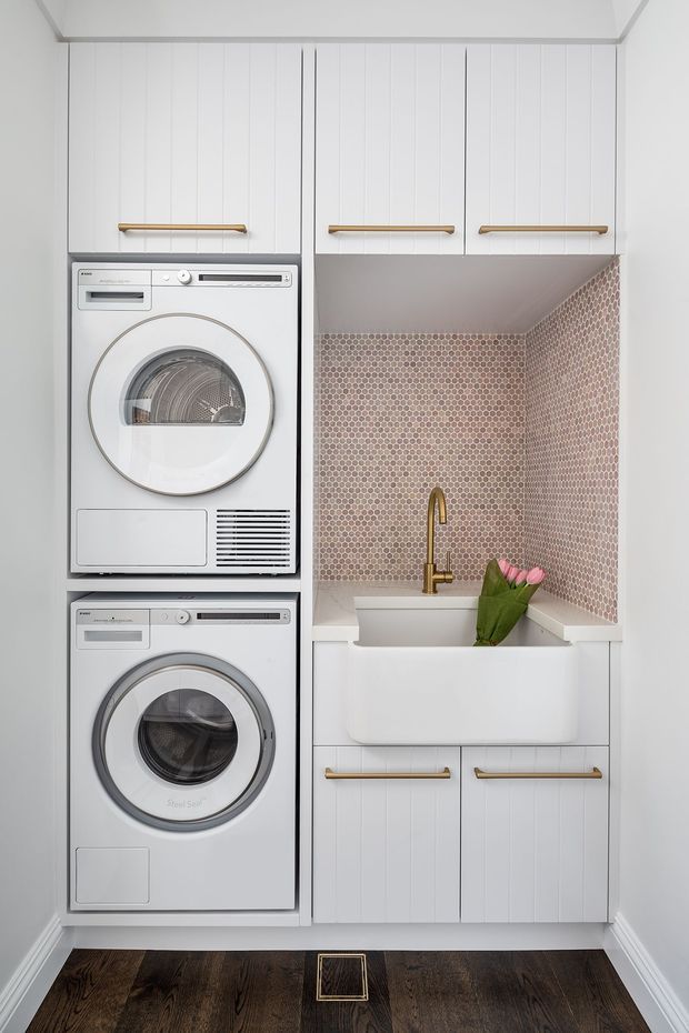 Compact, bright laundry room with stacked white front-loading washer and dryer, white vertical-panel cabinetry, brass bar handles and faucet, a deep white utility sink, and a pink-and-brown hexagonal tile splashback. Dark timber flooring and white trim complete the space.