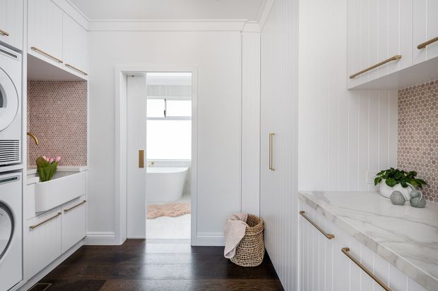 Bright, refined laundry room with white vertical-panel cabinetry, brass handles, a marble-look benchtop, and a dusty pink hexagonal tile splashback. Integrated laundry appliances are visible on the left, while a doorway opens to a bathroom with a freestanding bathtub. Dark timber flooring and soft decorative accents add contrast.
