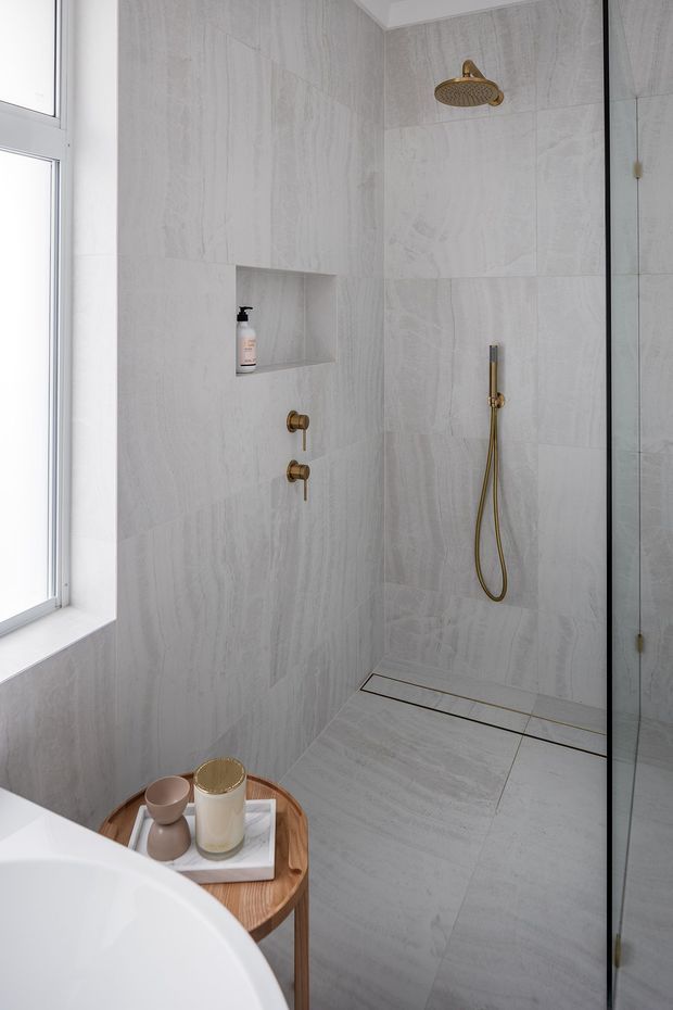 A bright, refined bathroom with a walk-in glass shower, large pale grey marble-look wall and floor tiles, a recessed shower niche, and brushed brass shower fixtures. A freestanding white bath and small timber side table with neutral accessories are visible in the foreground. Natural light enters through the tall window on the left.