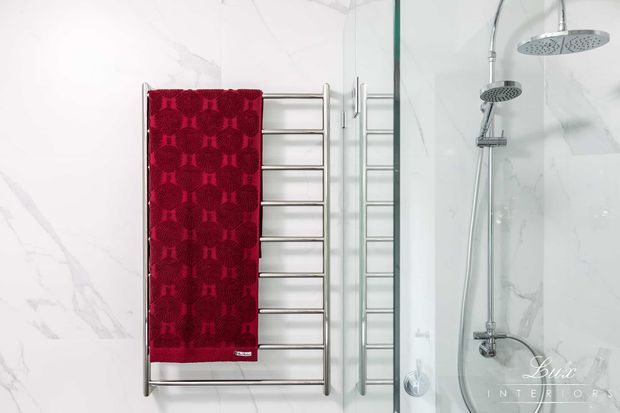 Bright contemporary bathroom detail featuring a chrome heated towel rail with a deep red towel, alongside a frameless glass shower enclosure with dual chrome showerheads and controls. The walls are clad in white marble-look surfaces with subtle grey veining.