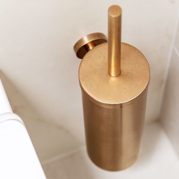 Close-up of a cylindrical brushed brass or bronze bathroom toilet brush holder with a matching vertical handle, positioned beside a white toilet against light neutral tiled walls.