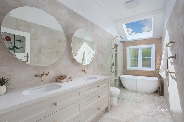 Bright, elegant bathroom with a long white double vanity, two round mirrors, wall-mounted brass taps, a freestanding bathtub, glass shower enclosure, toilet, skylight, and pale stone-look wall and floor finishes. A small stained-glass window is reflected in the mirror.