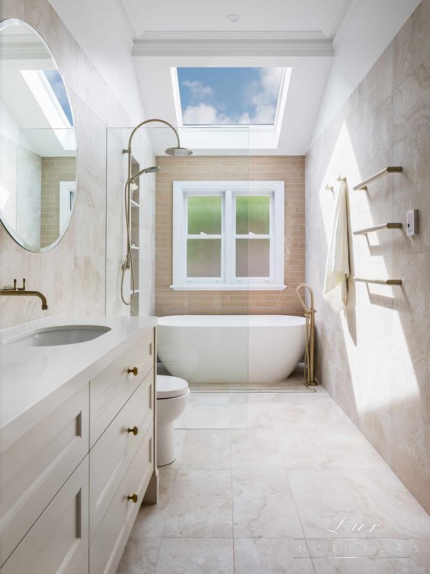 Bright, elegant traditional bathroom with a freestanding white bathtub beneath a skylight, a separate shower, white vanity cabinetry, pale stone-look wall and floor finishes, brass fixtures, a frosted-glass window, and a heated towel rail. The room has abundant natural light and refined classic detailing.