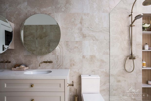 A refined bathroom featuring full-height beige and cream stone-look wall tiling, a pale grey vanity with integrated basin, round wall mirror, brass wall-mounted tapware, and a shower with matching brass fittings. A partially visible recessed shelving niche provides storage, while the overall design is softly traditional and elegant.