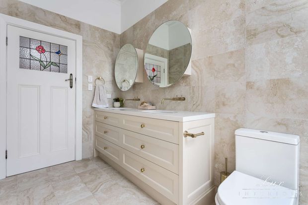A bright, elegant traditional bathroom featuring a large cream vanity with drawers, dual basins and brass hardware, marble-look stone wall and floor finishes, oval mirrors, a white toilet, and a decorative stained-glass door panel. The room has refined detailing and a warm neutral palette.