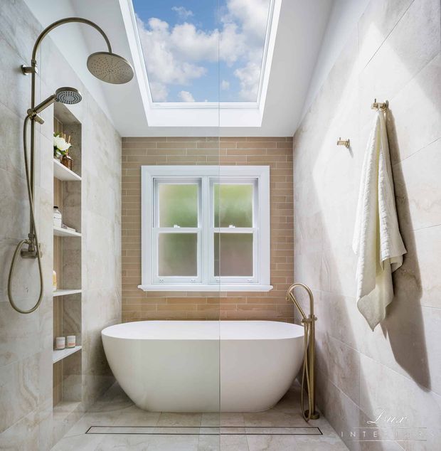 Bright, narrow bathroom with a freestanding oval bathtub beneath a window, a brass floor-mounted tap, a brass rainfall shower, built-in recessed shelving, pale stone-look wall and floor finishes, and a large skylight. White towels hang on brass hooks along the right wall.