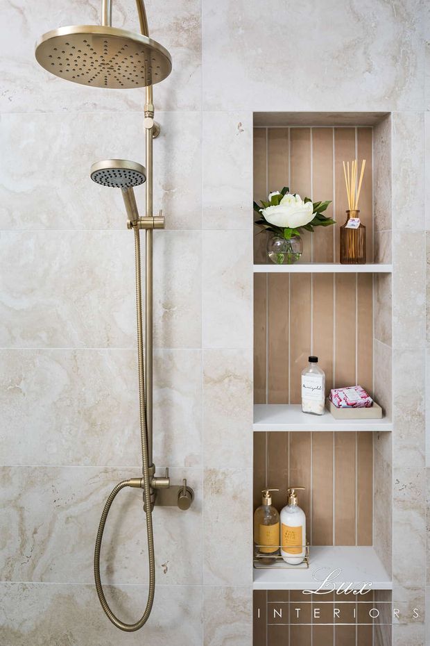 Close-up of a refined bathroom shower area with large pale stone-look wall tiles, brushed brass rainfall and handheld shower fittings, and a built-in recessed niche with multiple shelves. The niche has warm vertical tile detailing and contains toiletries, a reed diffuser, and a flower arrangement. The overall look is warm, polished, and luxurious.