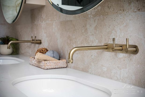 Close-up of a refined bathroom vanity featuring a white integrated basin, pale beige natural-stone wall finish, and brushed brass wall-mounted tapware. A rounded mirror, small plant, and woven basket with towels add warm decorative detail.