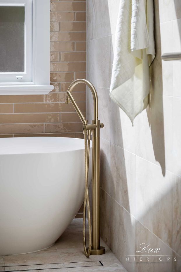 Close-up view of a refined bathroom featuring a freestanding white bathtub, brushed brass floor-mounted tub filler with handheld shower, warm beige subway wall tiles, pale stone-look floor tiles, a white-framed window, and a softly draped white towel. Natural light creates a calm, warm atmosphere.