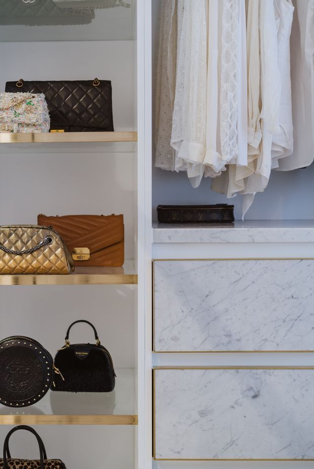 Close-up of a luxury walk-in wardrobe or dressing-room storage system. Open shelving displays handbags, while a rail holds white and cream garments. The built-in cabinetry features pale grey surfaces, light timber shelving, white marble-look drawer fronts with grey veining, and thin brass-gold trim.