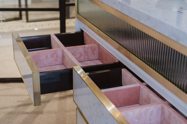 Close-up of a luxurious built-in cabinetry installation with multiple open drawers, pink and white marble-patterned drawer interiors and fronts, dark drawer boxes, brushed brass trim, and a vertically ribbed dark feature panel beneath a white marble benchtop. The image focuses on detailed fixed joinery rather than a complete room.