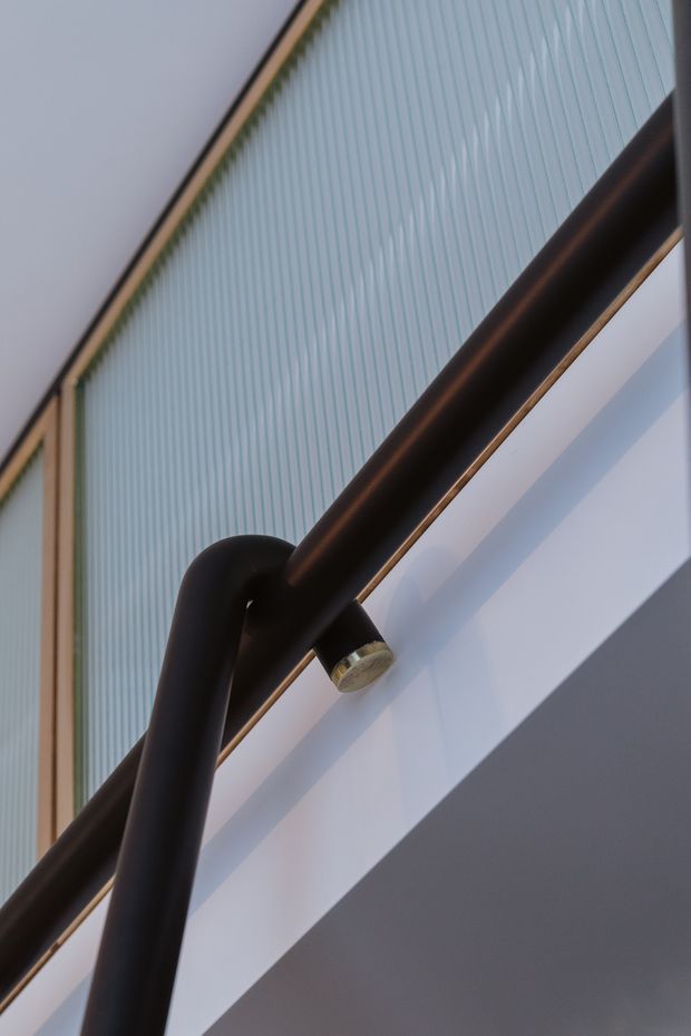 Close-up architectural detail of a dark tubular metal handrail with a curved joint and visible mounting detail, set against a pale blue vertically ribbed translucent or glazed wall and crisp white architectural surfaces. The tightly cropped upward perspective emphasizes the stair or balustrade structure and geometric lines.