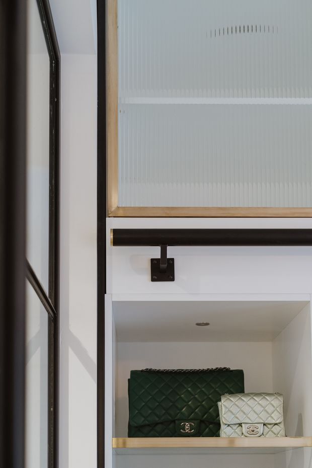Close-up architectural detail of a built-in display and storage unit. The composition shows white cabinetry with open shelving, pale timber shelf edges, black metal framing and hanging rail hardware, and a translucent ribbed glass panel above. Quilted handbags are displayed on the lower shelf, adding a dark green and cream accent.