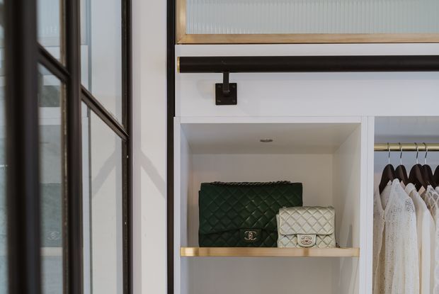 Close-up of a refined built-in wardrobe or dressing-room storage wall. White open shelving displays quilted handbags, with brass-toned shelf edging and dark metal hanging rails. A glazed black-framed door or partition is visible on the left, while white garments hang in the adjacent wardrobe section. The composition is highly detailed, with a restrained white, black, brass, and dark green palette.