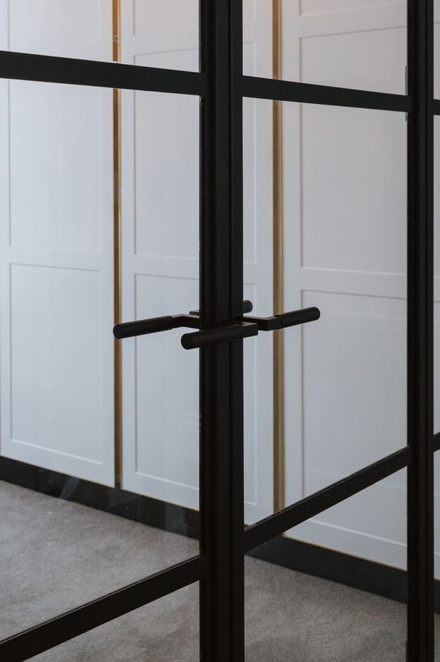 Close-up interior view of a black-framed glazed door or partition with rectangular metal mullions and horizontal rails, featuring a pair of slim black lever handles. White decorative wall panelling and pale flooring are visible behind the glazing, with dark skirting at the base.