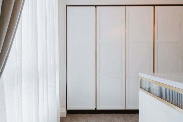 A bright, refined interior detail showing a wall of tall white paneled cabinetry or wardrobe doors with slim dark trim and warm metallic edging. Sheer white curtains and a heavier taupe curtain frame the left side, while the corner of a white built-in island or vanity with a fluted vertical feature and pale stone benchtop appears in the foreground. The space has a calm, minimalist contemporary character with soft natural light.