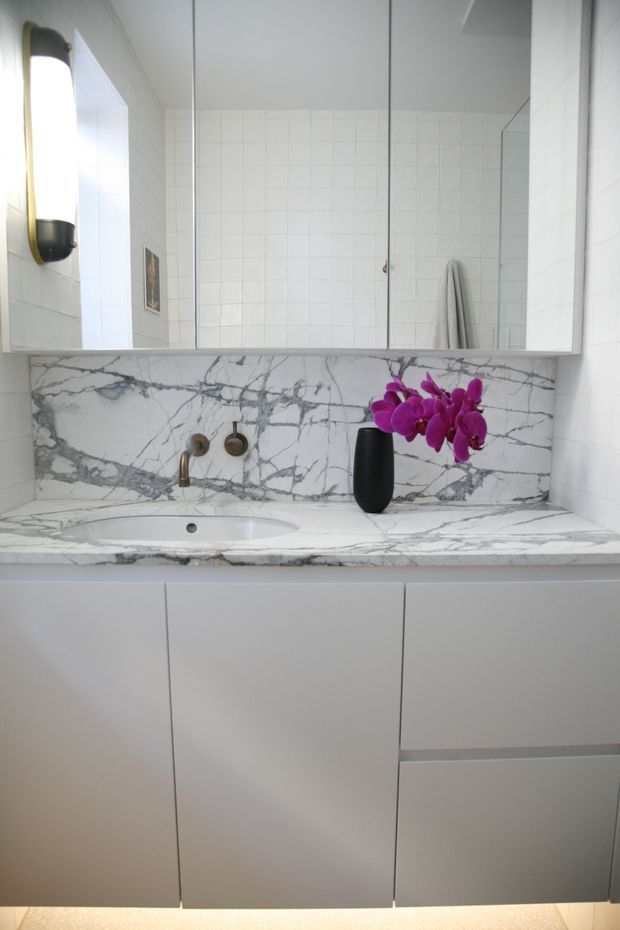 Bright contemporary bathroom vanity with a wide frameless mirrored cabinet, white tiled walls, white cabinetry, and a white marble-look stone benchtop and splashback with prominent grey veining. The vanity includes an integrated basin, wall-mounted bronze-toned tapware, and a small black vase with purple orchids.