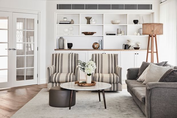 Bright, refined living room with white built-in shelving and cabinetry, styled with neutral ceramics and decor. Two striped upholstered armchairs face a round marble-topped coffee table, while a large grey sofa and woven floor lamp add layered texture. French doors and herringbone timber flooring contribute classic architectural character.