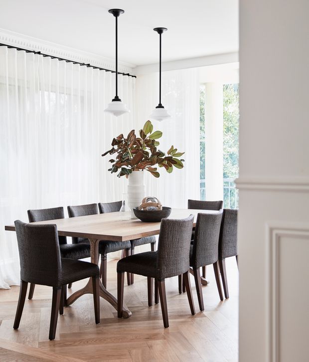 Bright, elegant dining room with a rectangular timber dining table surrounded by upholstered woven dining chairs. The room features sheer full-height curtains, large windows, white walls with decorative traditional moulding, pendant lights with black stems and frosted shades, and light timber flooring. A large leafy arrangement provides a natural focal point.