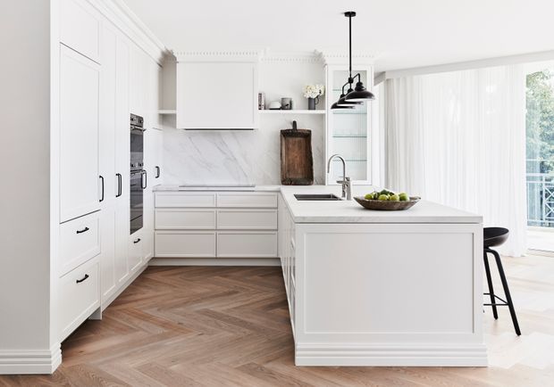 Bright, refined kitchen with white shaker-style cabinetry, integrated appliances, a large island with sink and seating, marble-look splashback, open shelving, black pendant lighting, and warm herringbone timber flooring. Floor-to-ceiling glazing and sheer curtains open toward a balcony or exterior view.