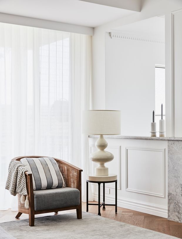 Bright, refined living room corner featuring a woven timber-framed armchair with striped cushion and throw, a small round side table with a sculptural lamp, sheer full-height curtains, decorative wall panelling, and a white fireplace or built-in cabinet with a pale stone mantel. The room has warm herringbone timber flooring and a soft neutral palette.