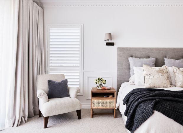 Bright, refined bedroom interior with a large upholstered bed, layered white and grey bedding, a dark knitted throw, bedside table, armchair, full-height curtains, plantation shutters, and decorative wall panelling with a cornice detail. The palette is soft and neutral with dark accents.