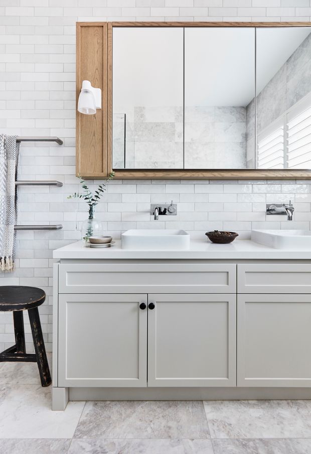 Bright, refined bathroom vanity with a double basin, white shaker-style cabinetry, pale stone-look floor tiles, white subway wall tiles, and a large timber-framed mirrored cabinet. Brushed metal wall-mounted taps, integrated lighting, towel rails, shutters, and restrained natural decor create a clean contemporary-traditional look.