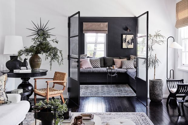 A stylish living room with a black-framed glazed partition enclosing a compact lounge or sitting area. The room features dark timber flooring, white walls, a black accent wall, a built-in dark sofa with patterned and blush cushions, woven seating, potted plants, framed artwork, layered rugs, and sculptural lighting. The overall look is refined, eclectic, and contemporary with strong black-and-white contrast and natural woven accents.