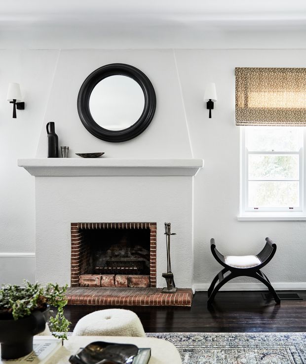 A refined living room centered on a traditional brick fireplace with a broad white mantel, circular black-framed mirror, and symmetrical black wall sconces. A small window with a patterned Roman blind sits to the right, while dark timber flooring, a black leather folding stool, a pale upholstered chair, and a patterned rug complete the room.