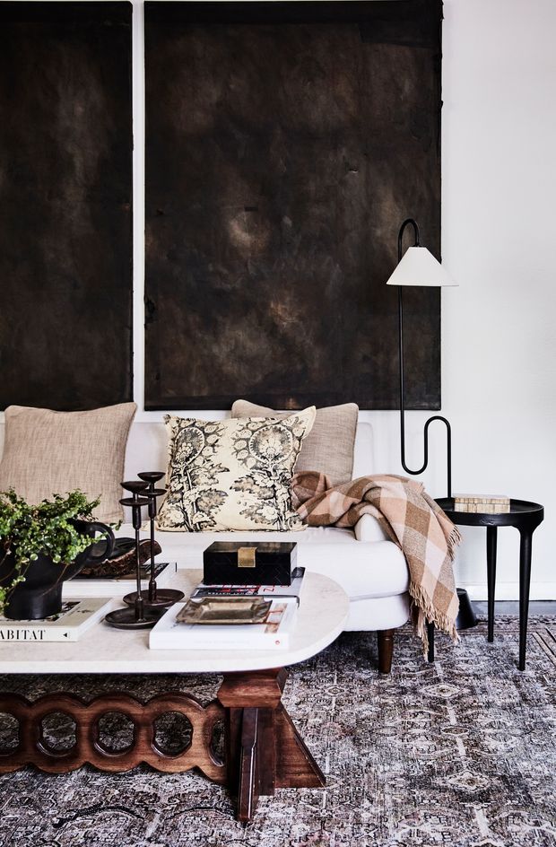 A styled living room vignette with a white sofa, layered patterned cushions, a brown checked throw, sculptural dark wood coffee table, black sculptural floor lamp, side table, potted greenery, and a large patterned rug. Two oversized dark abstract wall panels contrast with the white walls, creating a refined, moody, eclectic interior.