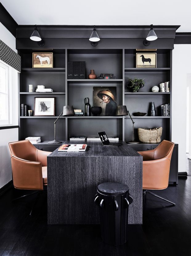 A sophisticated dark-toned home office with a built-in wall of charcoal shelving and cabinetry, displaying books, artwork and decorative objects. A substantial dark wood desk faces the shelving, accompanied by two tan leather chairs and a black stool. Black flooring, integrated task lighting, and monochromatic finishes create a refined, dramatic workspace.