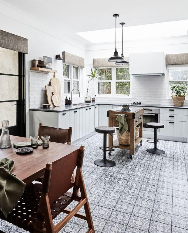 Bright, airy kitchen-dining space with white Shaker-style cabinetry, a dark benchtop, white subway-tile splashback, patterned tiled flooring, and black pendant lights. A central timber-topped island with stools sits in front of a bank of windows, while a timber dining table and leather-and-wood chairs occupy the foreground. Natural light enters through a large ceiling skylight and multiple windows.
