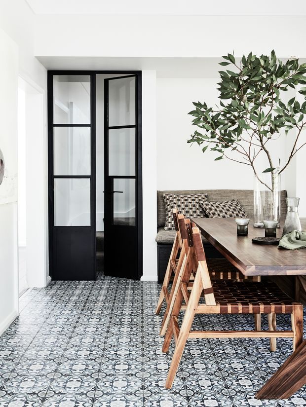 Bright, refined dining area with a long dark timber dining table, woven dining chairs, and a built-in patterned banquette. The room features decorative patterned floor tiles, crisp white walls and ceiling, and a black-framed glazed internal door leading to another space. A large indoor tree and neutral textured upholstery add an organic, contemporary character.