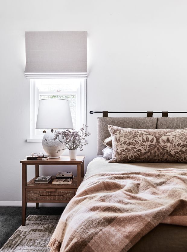 A softly styled bedroom with a partially visible bed, upholstered headboard, patterned neutral cushions, and a textured beige throw. A timber bedside table with woven drawer fronts, books, glasses, a ceramic vase, and dried floral branches sits beside the bed. The room has pale walls, a white-trimmed window with a Roman blind, and layered bedside lighting, creating a warm, calm, neutral interior.