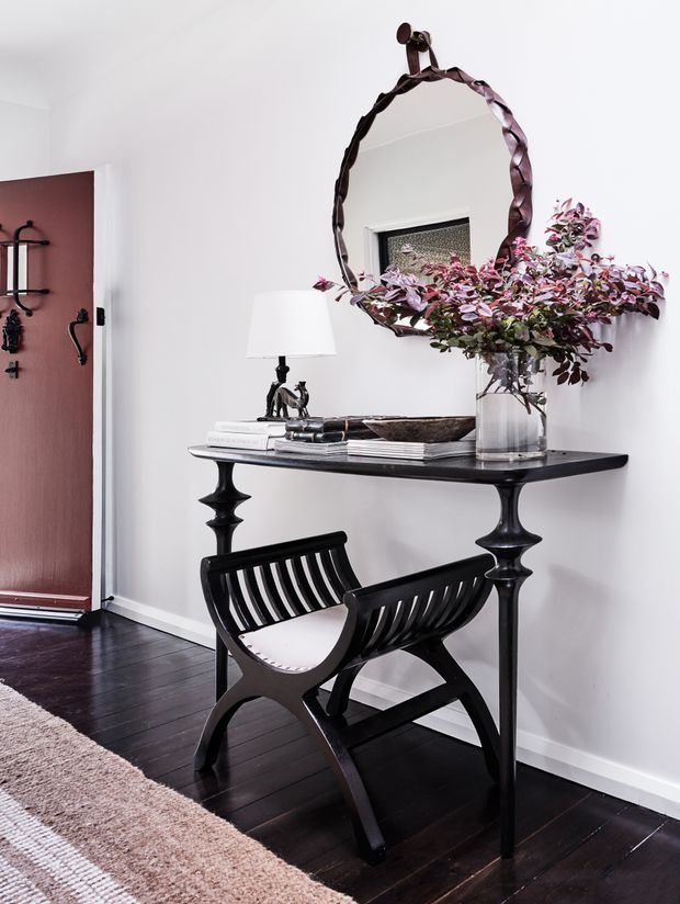 A styled entryway vignette with a black console table against a white wall, topped with books, a lamp, decorative objects, and a glass vase of leafy branches. A large irregular dark-framed mirror hangs above the console. A black sculptural chair sits in front, with dark timber flooring, a neutral rug, and a burgundy-red door visible to the left.