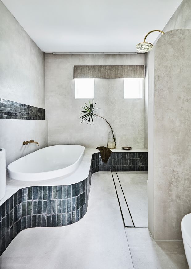 A refined contemporary bathroom with a sculptural freestanding white bathtub, curved built-in tiled platform, concrete-look walls and floor, dark vertically stacked tile accents, brass tapware, and a walk-in shower with a round overhead rain shower. A narrow textured window blind and sparse plant styling add warmth to the minimalist space.