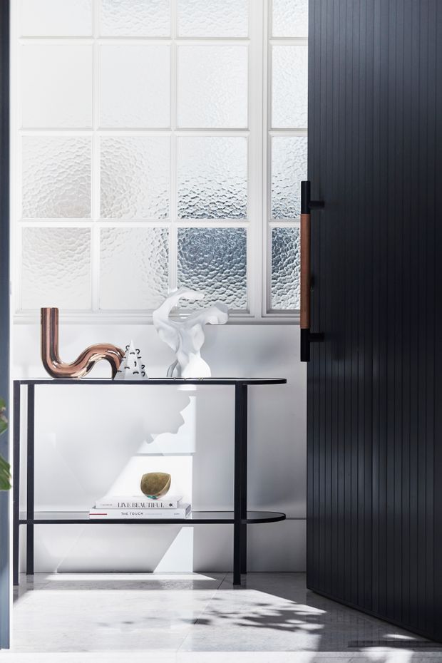 A bright, refined entryway vignette featuring a slim black metal console table with open shelving, sculptural white and copper decor, and a book. Behind it is a large grid of textured obscured-glass windows in white frames, while a tall dark vertically ribbed door or wall panel with a black handle occupies the right side. Strong sunlight creates crisp shadows across the pale stone or concrete floor.